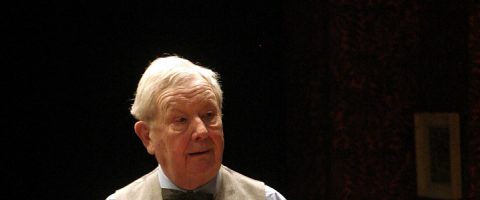 Valkohiuksinen iäkäs mies, jolla on vaalea paita, liivi ja rusetti, seisoo hämärässä huoneessa ja katsoo sivulle.