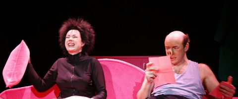 Kaksi näyttelijää istuu kirkkaan vaaleanpunaisella sohvalla; toinen, mustiin pukeutunut ja vaaleanpunainen tyyny kädessään, nauraa, kun taas toinen, toppapuvussa ja tekosian nenä päässä, lukee vaaleanpunaista lehteä vakavalla ilmeellä.