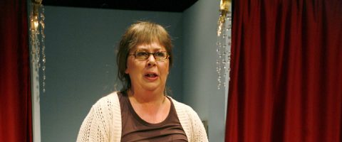 Silmälasipäinen nainen, jolla on lyhyet ruskeat hiukset, seisoo punaisten verhojen välissä, yllään beige villatakki ruskean paidan päällä, neutraalin sininen tausta ja roikkuvat kristallikoristeet molemmin puolin.