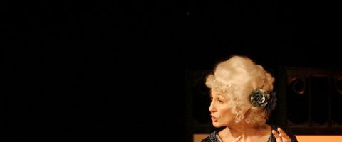 Vanhempi nainen, jolla on kiharat valkoiset hiukset ja jota koristaa suuri sininen kukka, puhuu intohimoisesti kasvot sivuttain tummaa taustaa vasten.