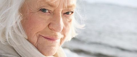 Lähikuva valkoiset hiukset ja vaaleat silmät omaavasta, vaalea huivi päässään olevasta iäkkäästä naisesta, joka hymyilee hellästi kameraan katsoen, taustan ollessa sumea.