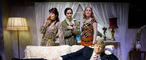 Neljä ihmistä vintage-tyylisessä olohuoneessa: kolme naista seisoo sohvan takana, yksi tupakoi, yksi pitää kissaa kädessään ja yksi poseeraa itsevarmasti; pukumies makaa sohvalla ja katsoo kameraan.