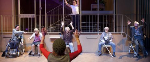 Ryhmä iäkkäitä aikuisia, joista osa on pyörätuolissa ja osa istumassa, nostaa kätensä ja katsoo kohti henkilöä, joka seisoo kädet ylhäällä edessä. Toinen henkilö seisoo kaiteen takana olevalla korokkeella kädet ylhäällä.