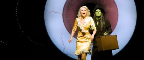 Kaksi naista seisoo näyttämöllä suuren, valaistun silmätaustan edessä; toisella on keltainen mekko ja hän näyttää yllättyneeltä, kun taas toinen, vihreäihoinen ja mustahattuinen nainen seisoo hänen takanaan matkalaukku kädessään.