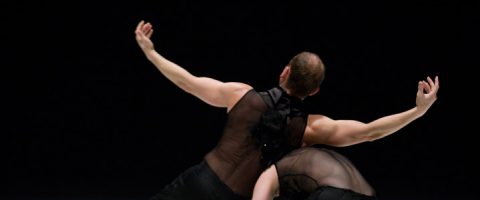 Kaksi tanssijaa mustissa puvuissa esiintyy lavalla tummaa taustaa vasten. Toinen tanssija kaartaa selkänsä ja ojentaa kätensä sirosti, kun taas toinen nojaa eteenpäin, osittain peitettynä.