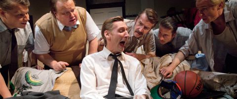 Kuusi miestä kerääntyy tiiviisti sängyn ympärille ja katselee keskittyneesti valkoiseen paitaan ja solmioon pukeutunutta miestä, joka näyttää huutavan tai nauravan. Sängyllä näkyy koripallo ja vaatteita. Kohtaus näyttää dramaattiselta ja teatraaliselta.