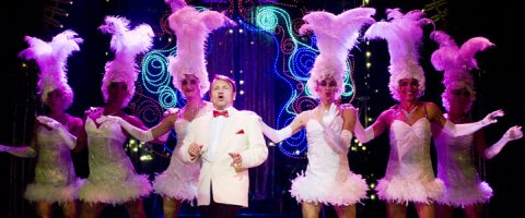Valkoiseen pukuun ja punaiseen rusettisolmioon pukeutunut mies laulaa lavalla kuuden tanssijan ympäröimänä, joilla on kimaltelevia valkoisia pukuja ja korkeita höyhenpäähineitä, ja taustalla on värikkäitä valoja ja koristeita.