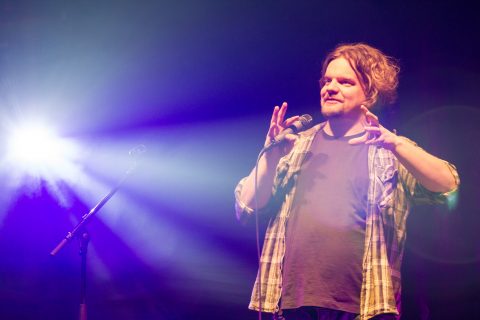 Mies seisoo mikrofoni kädessään elehtien käsillään. Taustalla on sininen näyttämövalaistus.