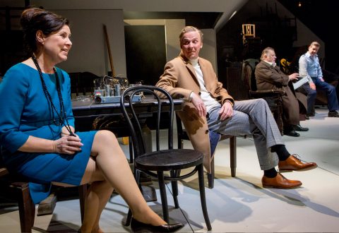 Neljä ihmistä istuu näyttämöllä dramaattisessa teatterikohtauksessa. Etualalla istuu nainen, jolla on tiilenpunainen mekko, ja kauempana takana istuu kolme pukuihin ja rentoihin vaatteisiin pukeutunutta miestä keskustelemassa, ja heidän ympärillään on rekvisiittaa ja huonekaluja.