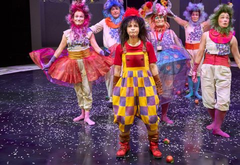 Joukko esiintyjiä värikkäissä, omituisissa puvuissa ja peruukissa seisoo konfettien peittämällä lavalla. Edessä olevalla henkilöllä on punainen paita ja kelta-violetti ruudullinen haalari, ja hän näyttää vakavalta, kun taas muut takana olevat näyttävät iloisilta.