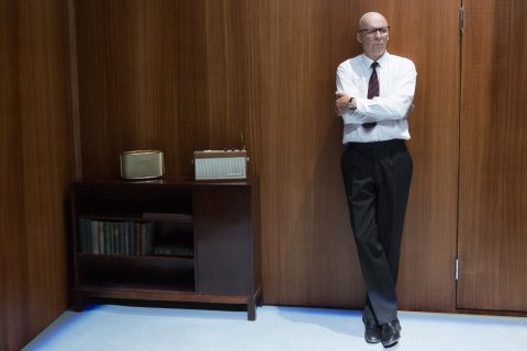 Valkoiseen paitaan ja solmioon pukeutunut mies seisoo kädet ristissä ja nojaa puuseinään, joka on radiota ja kirjoja sisältävän kaapin vieressä. Huoneessa on minimalistinen retrotyyli.