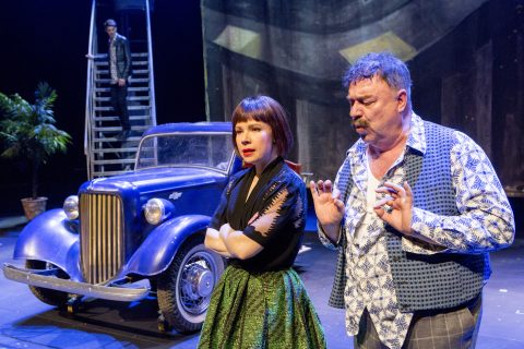 Kaksi näyttelijää esiintyy lavalla vanhan sinisen auton edessä; toinen seisoo kädet ristissä, toinen elehtii käsillään. Taustalla kolmas henkilö seisoo tikkailla ruukkukasvin vieressä.