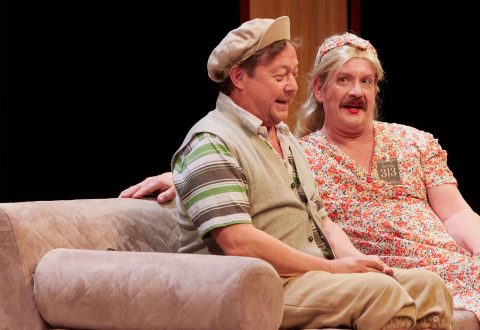 Kaksi miestä istuu näyttämöllä beigen sävyisellä sohvalla; toisella on lippis, raidallinen paita ja liivi, kun taas toisella on kukkamekko, vaalea peruukki ja punainen huulipuna, ja he osallistuvat humoristiseen teatteriesitykseen.