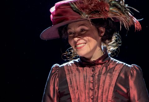 Nainen, jolla on vanha, tummanpunainen mekko ja suuri, koristeellinen ja höyhenillä koristeltu hattu, hymyilee näyttämövalaistuksessa, mikä viittaa teatteriesitykseen.