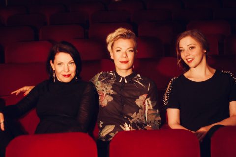 Kolme naista istuu punaisissa teatteripenkeissä ja hymyilee kameralle. Tausta on pehmeästi valaistu ja epätarkka, ja heidän takanaan on tyhjiä istuimia. Jokaisella naisella on tummat tai kuviolliset vaatteet.