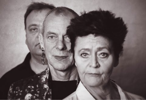 Kolme aikuista seisoo tiiviisti rivissä kameraan päin. He ovat asettuneet niin, että kukin henkilö peittää osittain takanaan olevan henkilön. Kuva on mustavalkoinen, ja ilmeet ovat vakavia.