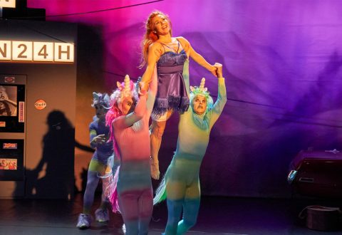 Kaksi sateenkaarenvärisiin yksisarvispukuihin pukeutunutta esiintyjää nostaa violettiin mekkoon pukeutunutta naista värikkäällä näyttämöllä, ja taustalla on toinen yksisarvinen esiintyjä ja kyltti, jossa lukee 24H.