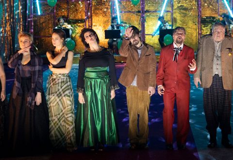 Kuusi värikkäisiin teatteripukuihin pukeutunutta ihmistä seisoo lavalla rivissä dramaattisin ilmein. Heidän takanaan muusikot soittavat kullanhohtoisen taustan ja vihreiden ilmapallojen edessä. Siniset valot lisäävät juhlallista tunnelmaa.