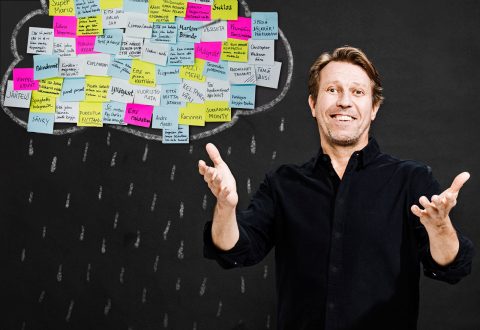 Hymyilevä mies seisoo liitutaulun edessä, hänen päänsä yläpuolella on pilvi värikkäitä tarralappuja ja pilvestä putoaa liidulla piirrettyä sadetta, aivan kuin hän toivottaisi sateen tervetulleeksi avosylin.