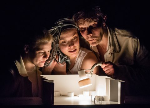 Kolme ihmistä tutkii keskittyneesti pientä arkkitehtonista pienoismallia, jota valaisee pöytälamppu muuten pimeässä ympäristössä. Heidän kasvonsa ovat dramaattisesti valaistut, kun he nojaavat tiiviisti yhteen mallin yli.