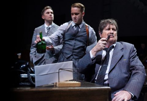 Lavalla on kolme pukumiestä. Yksi istuu pöydän ääressä piippua polttaen, ja kaksi muuta seisovat hänen takanaan; toisella on kädessään vihreä pullo. Kohtauksessa on dramaattinen, vintage-tunnelma.