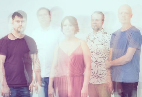 Viisi ihmistä seisoo rivissä vaaleaa taustaa vasten. Kuva on tarkoituksellisesti epätarkka ja siinä on kaksoisvalotus, mikä antaa hahmoille aavemaisen, läpinäkyvän vaikutelman. Heillä on yllään vaaleanväriset rennot vaatteet.