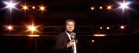 Pukumies seisoo lavalla mikrofoni kädessään ja katsoo luottavaisesti kameraan. Hänen takanaan loistavat kirkkaat lavavalot, jotka luovat dramaattisen tunnelman.