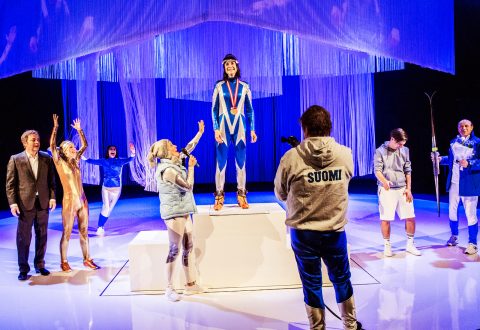 Sinivalkoiseen pukuun pukeutunut henkilö seisoo korokkeella mitali kädessään ja hymyilee. Toiset, jotkut urheiluasuissa, hurraavat, juhlivat ja ottavat valokuvia näyttämön dramaattisen valaistuksen ja ripustettujen koristeiden alla.