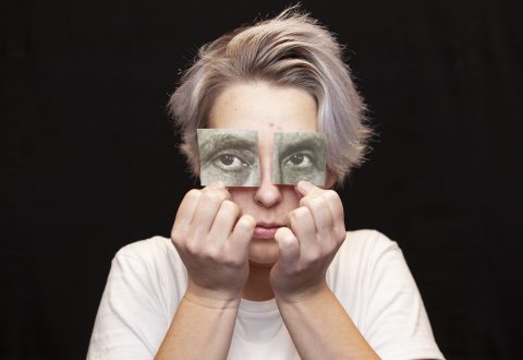 Henkilö, jolla on lyhyet vaaleat hiukset, pitää kahta korttia, joissa on piirretyt silmät, omien silmiensä päällä, mikä luo surrealistisen vaikutelman. Hänellä on valkoinen paita ja hän seisoo mustaa taustaa vasten.