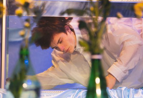 Valkoiseen paitaan pukeutunut nuori henkilö on upotettu lasiseen vesisäiliöön ja vaikuttaa mietteliäältä tai pohdiskelevalta. Etualalla on vihreitä lasipulloja ja keltaisia kukkia.