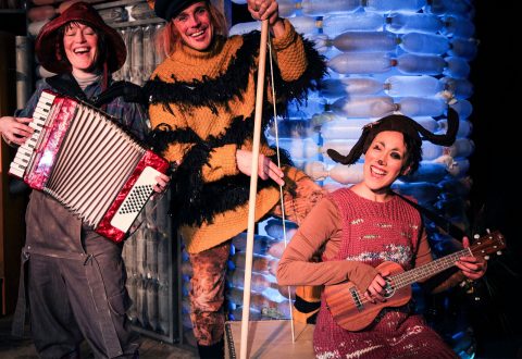 Kolme hassuihin pukuihin pukeutunutta ihmistä poseeraa leikkisästi sinisen taustan edessä. Yksi soittaa harmonikkaa, toisella on kelta-musta mehiläispuku, ja kolmas, jolla on antennit, soittaa pientä kitaraa.