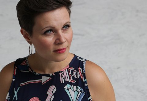 Nainen, jolla on lyhyet ruskeat hiukset ja rengaskorvakorut, katsoo hieman ylöspäin. Hänellä on yllään hihaton mekko, jossa on värikkäitä geometrisia kuvioita ja kankaassa näkyvät kirjaimet ME. Tausta on vaalea ja yksivärinen.