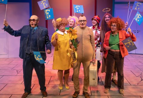 Joukko värikkäisiin, omituisiin asuihin pukeutuneita ihmisiä seisoo yhdessä sisätiloissa hymyillen ja pitäen kädessään sinisiä lippuja, joissa on keltainen kysymysmerkki. Yksi ruskehtavaan bodyyn pukeutunut henkilö pitelee kukkia ja matkalaukkua. Kohtaus näyttää teatraaliselta ja iloiselta.