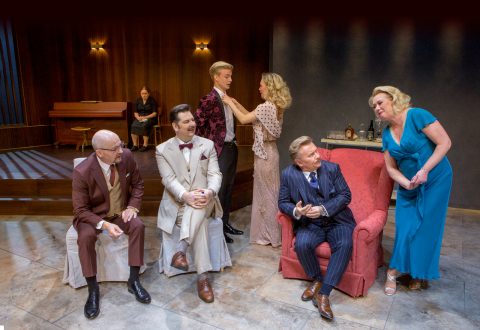 Lavalla on kuusi juhlapukuista ihmistä; kolme miestä istuu ja puhuu, heidän vieressään seisoo sinipukuinen nainen, ja kaksi ihmistä on vuorovaikutuksessa taustalla. Ympäristö on tyylikäs huone, jossa on piano.