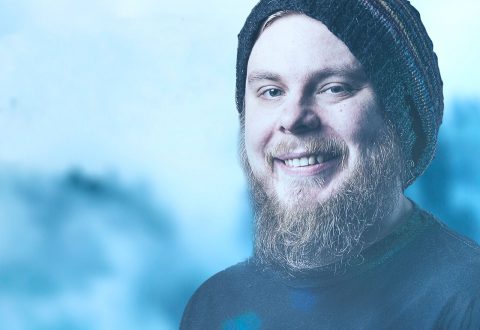 Parrakas mies, jolla on neulottu pipo ja tumma paita, hymyilee kameralle pehmeän sinistä sumeaa taustaa vasten.