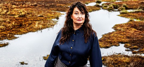 Nainen, jolla on pitkät, ruskeat, aaltoilevat hiukset ja tumma pusero, seisoo rämeisen maiseman edessä, jossa on vesilaikkuja ja ruskeita ruohoja.