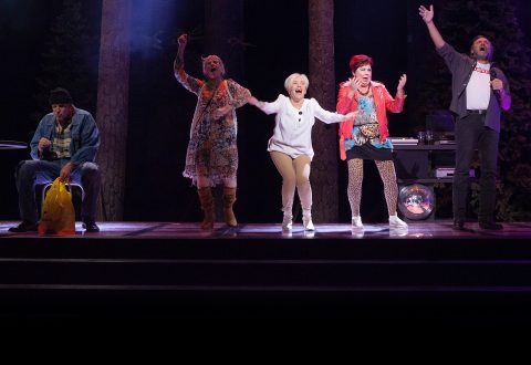 Viisi aikuista seisoo lavalla puiden rekvisiitan edessä; neljä laulaa tai elehtii energisesti, kun taas yksi istuu vasemmalla keltainen ostoskassi kädessään ja katsoo alaspäin. Näyttämön valaistus valaisee esiintyjät.