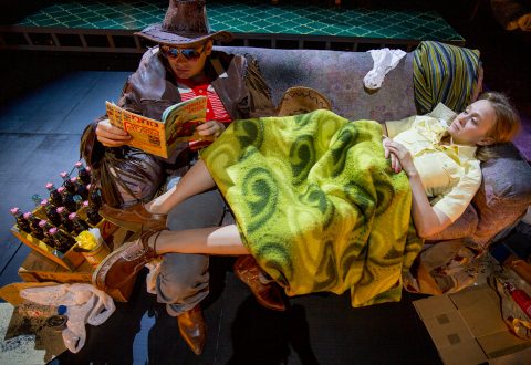 Cowboy-hattuinen ja aurinkolaseja käyttävä henkilö lukee sarjakuvaa sohvalla toisen vihreän peiton alla makaavan henkilön vieressä, ja lattialla on pulloja ja laatikoita teatteriympäristössä.