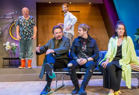 Lavasteissa on viisi ihmistä, kolme miestä ja kaksi naista. Kaksi miestä ja yksi nainen istuvat sohvalla, ja kaksi miestä seisoo heidän takanaan. Lavastukseen kuuluu polkupyörä, kirkas sisustus ja rento vaatetus.