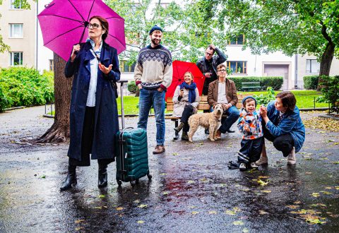 Nainen vaaleanpunaisen sateenvarjon ja matkalaukun kanssa seisoo puistonpenkin vieressä, kun hänen takanaan on joukko ihmisiä ja koira. Kaksi aikuista pitelee sateenvarjoja, ja lapsi hymyilee, kun toinen henkilö kyykistyy ottamaan valokuvaa. Maa on lehtien kastelemaa.