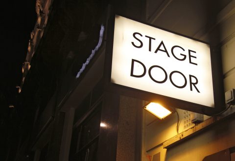 Valaistu STAGE DOOR -kyltti on asennettu rakennuksen ulkoseinään yöllä, ja se valaisee sisäänkäynnin alapuolella.