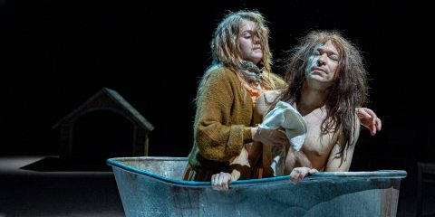 Vanhanaikaisiin vaatteisiin pukeutunut nainen auttaa pitkätukkaista, paidatonta miestä kylpemään lavalla olevassa metallisessa ammeessa, jonka hämärässä taustalla näkyy koirankoppi.