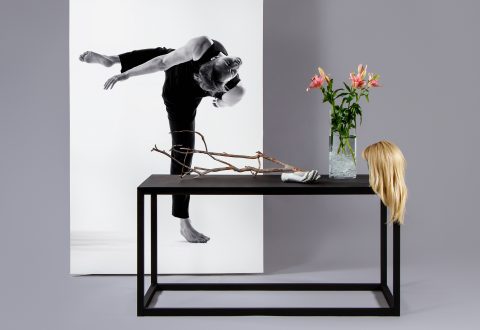 Minimalistinen kohtaus, jossa mustalla pöydällä on lasimaljakko, jossa on vaaleanpunaisia liljoja, puun oksa ja mannekiinikäsi. Pöydällä roikkuu vaalea peruukki, ja sen takana on mustavalkoinen valokuva liikkeessä olevasta tanssijasta.