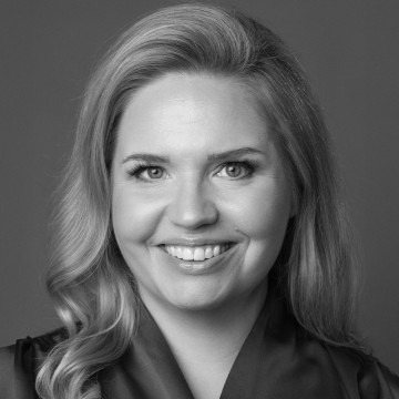 Nainen, jolla on pitkät, aaltoilevat hiukset, hymyilee kameralle. Hänellä on vaaleat silmät ja yllään tumma toppi. Kuva on mustavalkoinen ja siinä on tavallinen tausta.