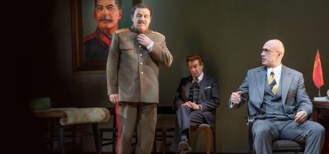 Kolme miestä huoneessa: yksi neuvostoliittolaiseen sotilaspukuun pukeutunut mies seisoo Stalinin muotokuvan vieressä, toinen istuu puvussaan ja katsoo alaspäin, ja kolmas kalju mies, jolla on silmälasit ja puku, tekee eleitä istuessaan pöydän ääressä, jossa on pieni Neuvostoliiton lippu.