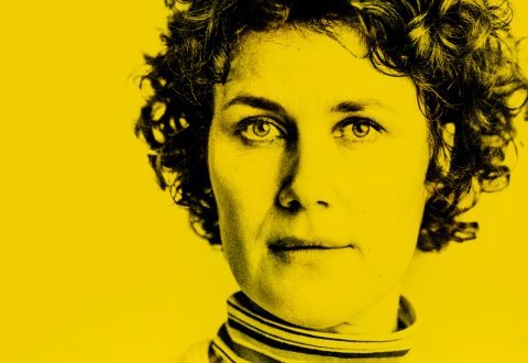 Lähikuva henkilöstä, jolla on kiharat hiukset ja vakava ilme, ja joka on esitetty voimakkaassa kontrastissa keltaisella värillä. Henkilöllä on yllään raidallinen poolopusero.
