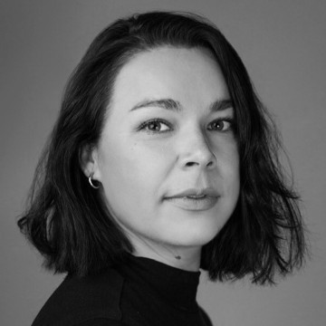 Nainen, jolla on tummat, olkapäille ulottuvat hiukset ja vaaleat silmät, katsoo kameraan. Hänellä on yllään tumma paita ja pienet rengaskorvakorut. Kuva on mustavalkoinen, ja sen tausta on neutraali.