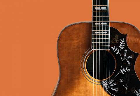Lähikuva akustisesta kitarasta oranssia taustaa vasten, jossa näkyy osa rungosta, koristeellinen, kukkakuvioinen soittimen suojus ja kaulan alaosa, jossa on näkyvissä jouset ja kielet.