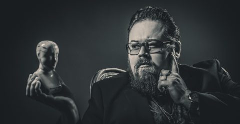 Silmälasipäinen, parrakas, pukuun pukeutunut mies istuu mietteliäänä pitelemässä keraamista frenologiapäätä toisessa kädessä ja koskettelemassa kasvojaan toisella kädellä. Kuva on mustavalkoinen.