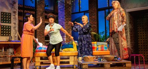 Neljä naista näyttelee tarmokkaasti värikkäällä näyttämöllä, jossa on tiiliseinät, suuret ikkunat ja trooppinen sisustus. Yhdellä naisella on päällään kypärä ja urheiluvaatteet, kun taas muut ovat pukeutuneet eläväisiin, rentoihin asuihin.